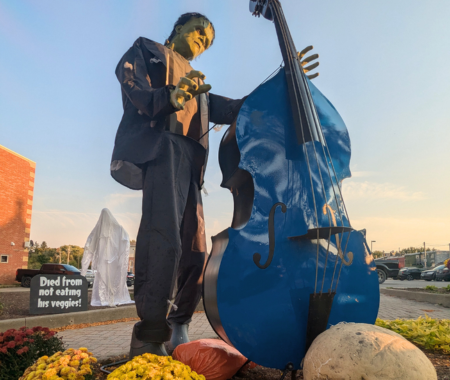 Statue of a green skinned spooky man stands next to a large blue bass like he is strumming it.